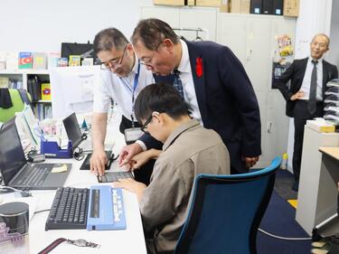 盲ろう職員の勤務状況の見学の様子。職員の使用する点字シートや点字ディスプレイを増子事務次官が触っている。