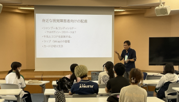 小林教授が「身近な視覚障害者向けの配慮」というタイトルのスライドを投影しながら説明しており、参加者と本学学生たちが聞いている。写真には7名ほどの聴講者の後ろ姿が写っている