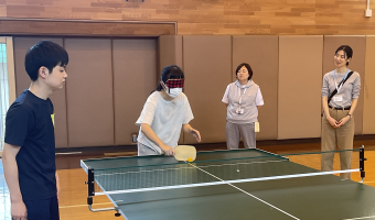 参加者の1人がアイマスクをして、サウンドテーブルテニスの体験でこちら側に玉を打ち換えそうとしている。卓球台の横に本学学生が立っており、参加者の広報では様子を見ているスタッフが2名立って様子を見ている