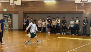 参加者の1人がアイマスクをしてサッカーボールを蹴ろうとしている。横に本学学生が立って教えており、壁沿いに並んで立っている他の参加者や学生らが15名ほどこちらを見ている